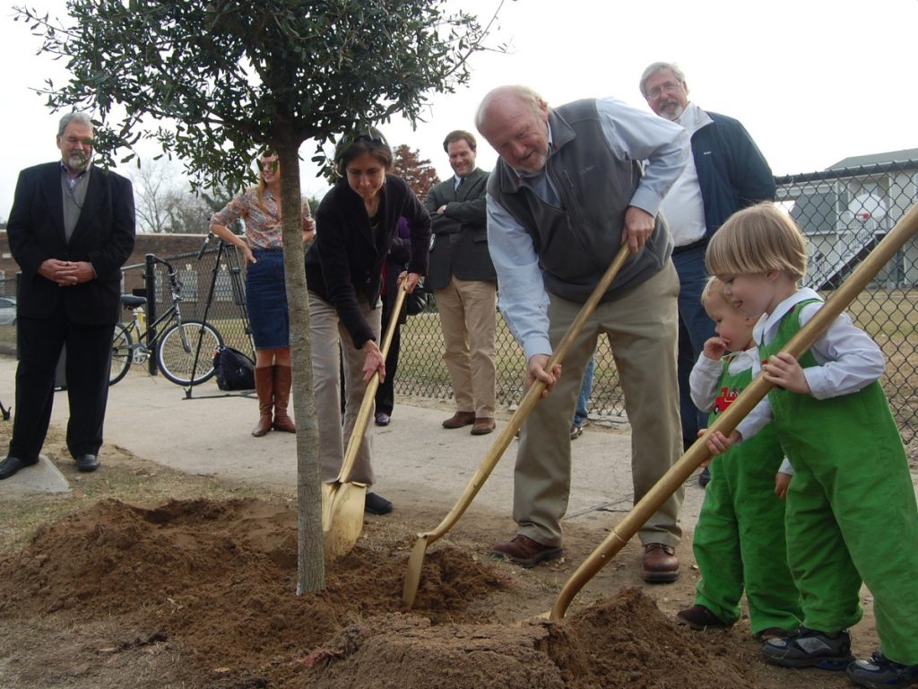 A group of people ceremoniously planting a tree, including well dressed business people and children dressed in green.