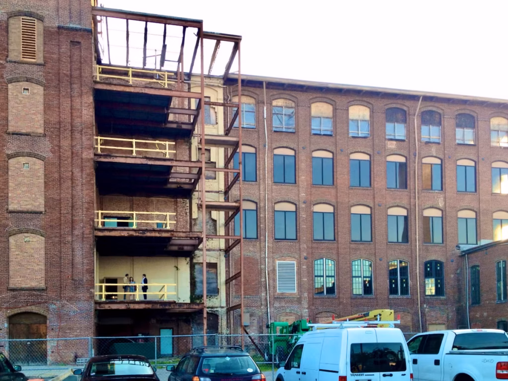 Brick and arched-window facade of the Cigar Factory with exposed exterior steel frames, partial scaffolding, and parked vans below.