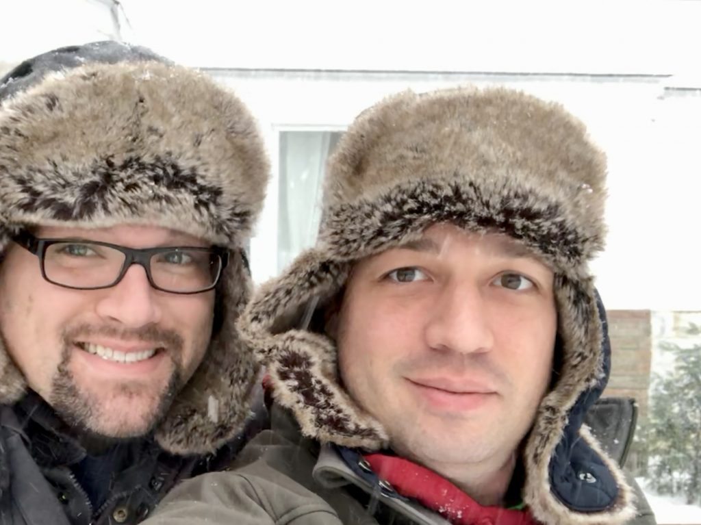 Bryan McElveen and his husband Adam dressed in winter attire with a snowy backdrop behind them.