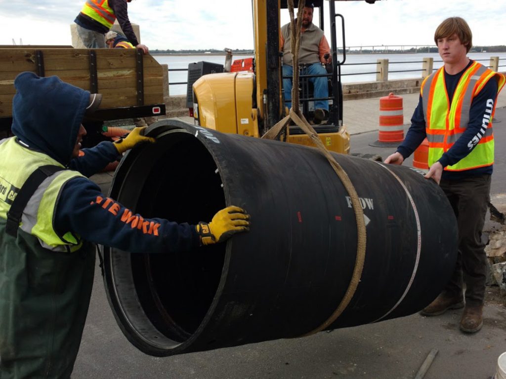 Workers handling a large black pipe containing a check valve, preparing to install it under a street in Charleston.