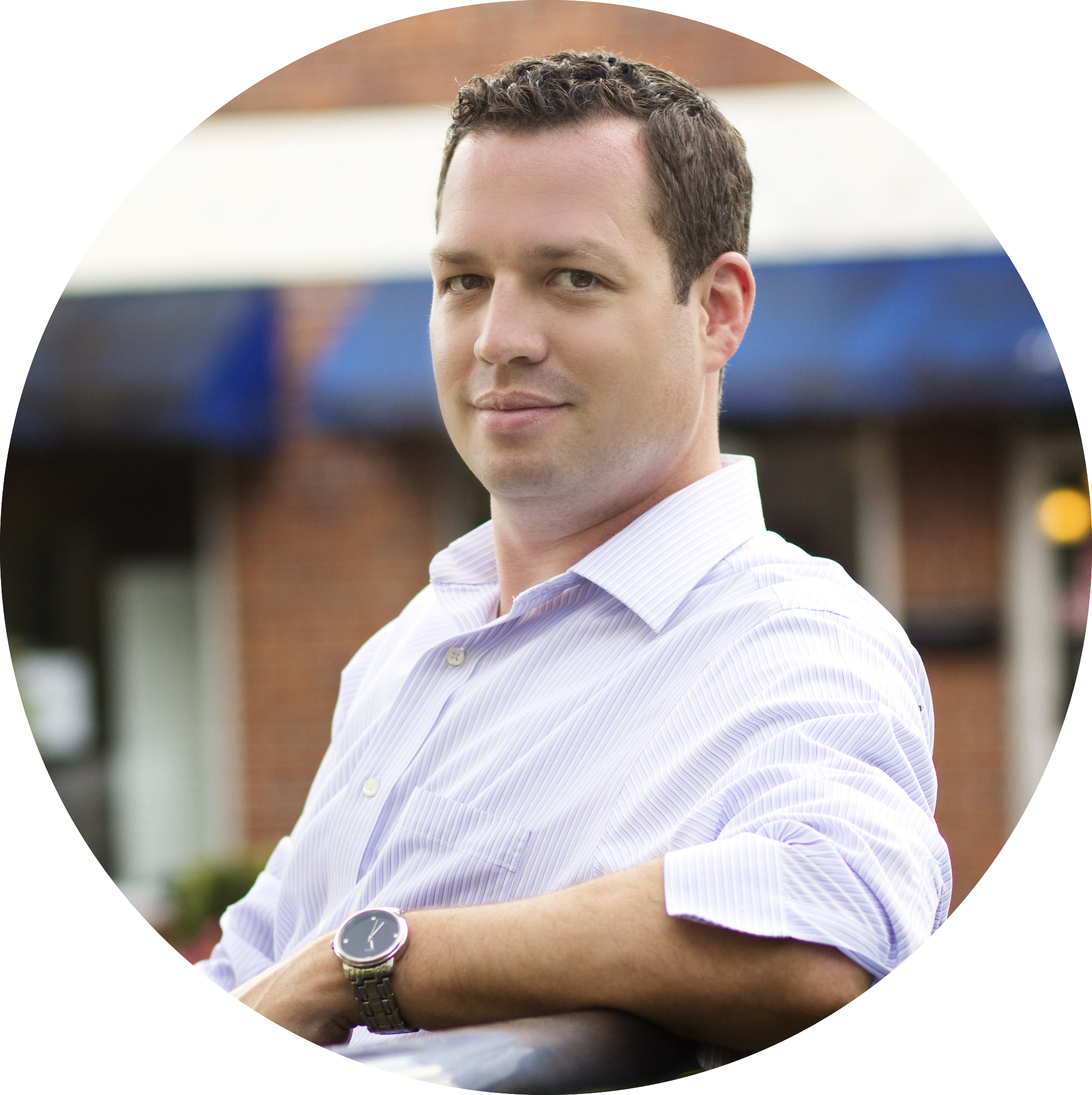 A circular cropped photo of real estate broker Bryan McElveen looking to his left. He has short brown hair, a longsleeve collared pink shirt with rolled up sleeves, and a silver wristwatch.