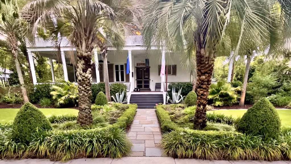 Front walkway leading to a white Charleston-style home entrance, framed by tall palm trees, manicured shrubs, and neatly trimmed hedges in bright greenery.