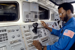 Astronaut Ron McNair in the cockpit of the Space Shuttle, seated at the control console and operating switches, with the instrument panel and hand controller in the foreground.