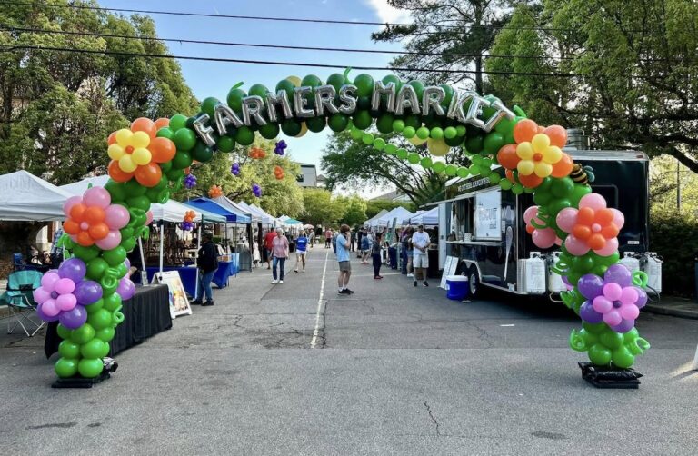 Outdoor Summerville Farmers Market entrance with an arch made of green, orange, pink, and purple balloons over a driveway lined with vendor tents and shoppers.