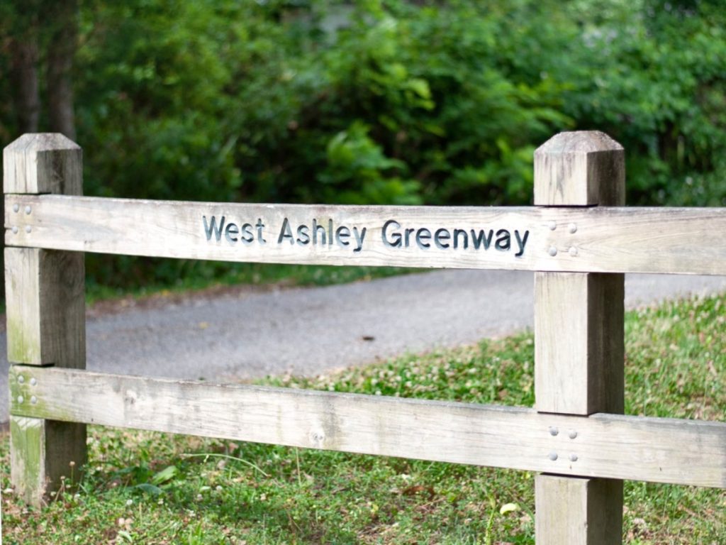 A wooden fence with the signage "West Ashley Greenway" with wild green shrubbery in the background.