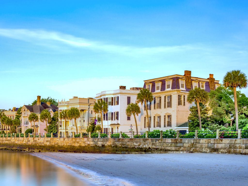 Large homes positioned close together along the seawall of The Battery with palmetto trees and other greenery around them. The view is from the small beach in front of The Battery with a small bit of water that is lightly reflecting the homes above it.
