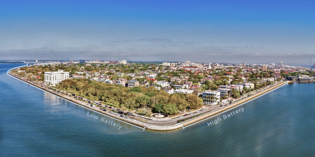 An aerial view of the southern Charleston peninsula from the above the blue water of Charleston Harbor, with a vantage point that appears triangular. The image is annotated to indicate the High Battery along the east side and the Low Battery on the south side.