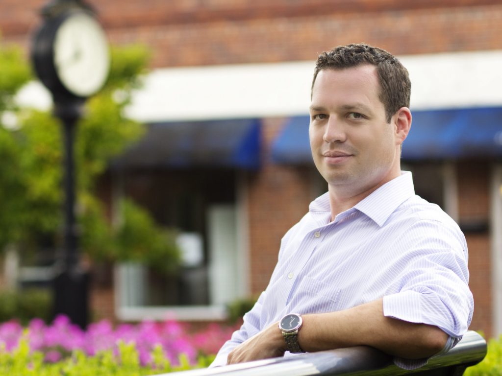 Bryan seated on a park bench wearing a light colored shirt and a wristwatch, looking towards the camera with a large street clock in the background.