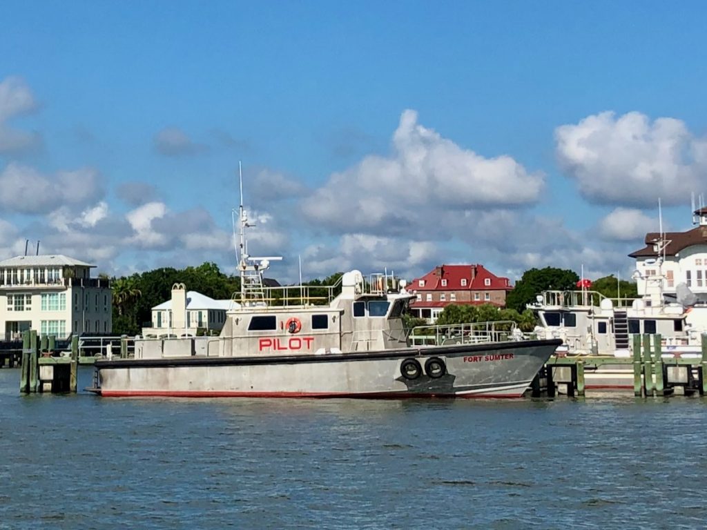 A view from the blue water of Charleston Harbor of a few boats moored to a dock, featuring a gray boat with the labels "PILOT" and "Fort Sumter" printed in red lettering. There are historic homes and boat facilities in the background amongst green trees.