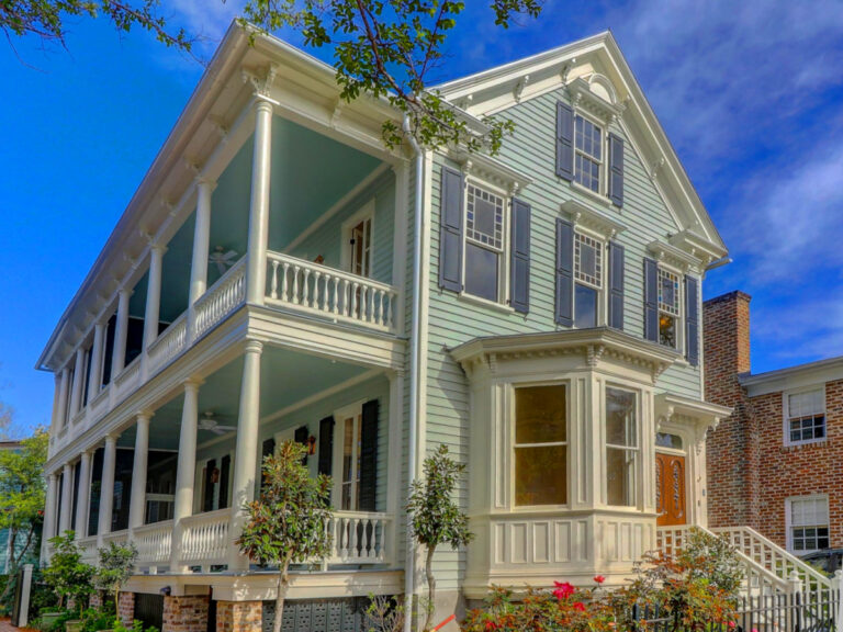 Front exterior of a two-story green house with white columns, covered porch, and blue shutters under a bright sky.