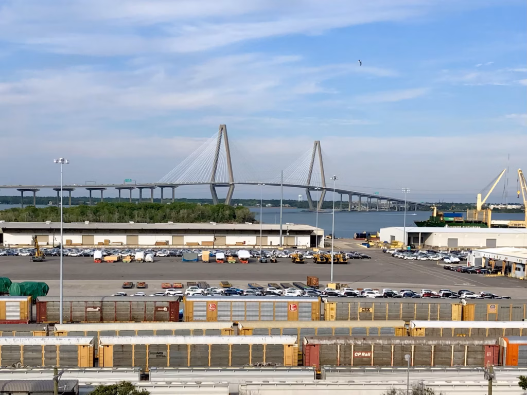 A busy waterfront shipping terminal with layers of trains and freight, with the Ravenel suspension bridge spanning the background under a partly cloudy sky.