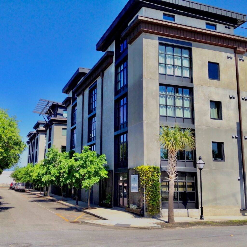 A street view of the condominium building One Cool Blow – the concrete and metal structure fill most of the view, lined by green street trees under blue sky.