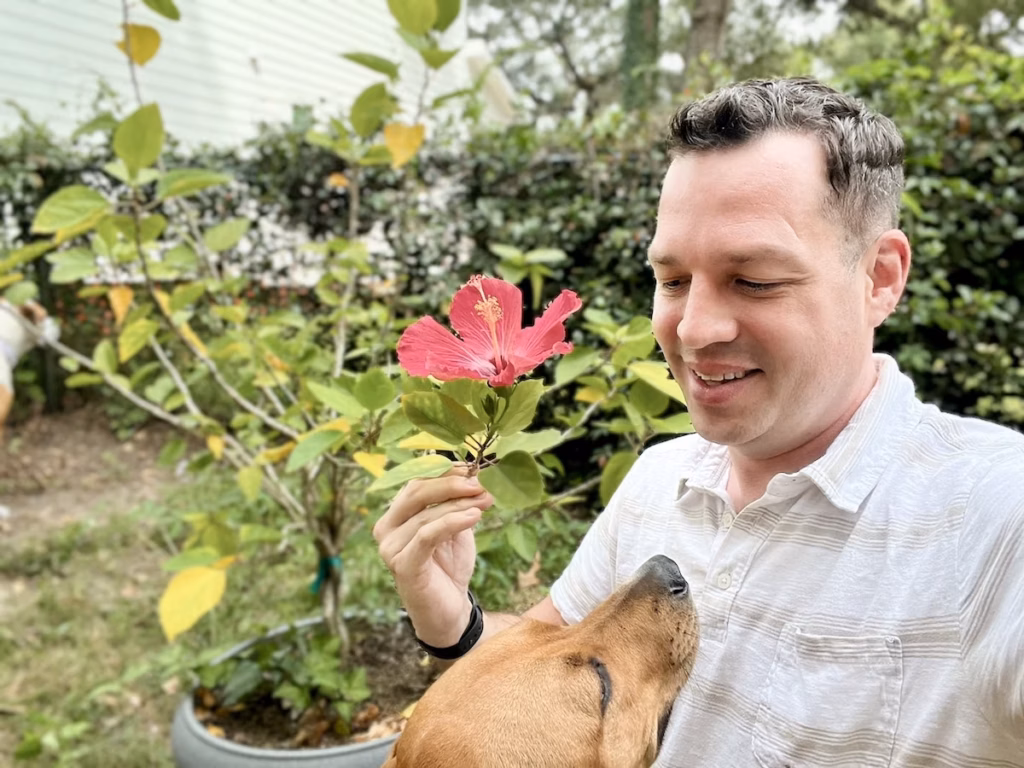 Bryan in a garden holding a small flower near his face while a tan dog sits beside him among green shrubs and trees.