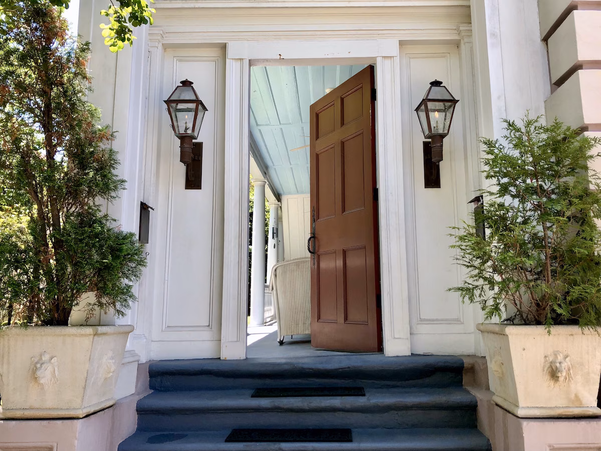 An open wooden door in the white exterior siding of a Charleston Single House, flanked by two dark metal lanterns and large green plants in square white planter pots.