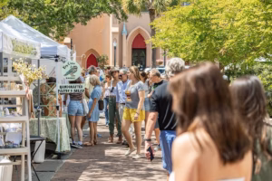 Outdoor farmers market walkway with vendor tents and shoppers passing by a storefront with arched doorways and red accents under bright, leafy trees.
