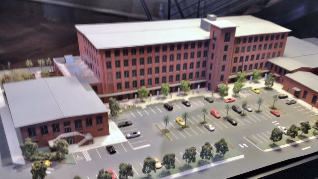 Scale model of a red-brick industrial building with a central parking lot, small trees, and miniature cars under an overhang.