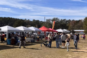 Outdoor farmers market on a grassy field with rows of white canopy tents, shoppers and families walking between booths under a blue sky.