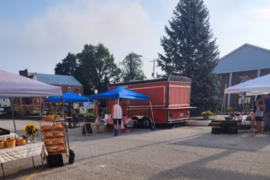 Outdoor farmers market with canopy tents, vendor tables, and a red delivery truck parked along a town street.