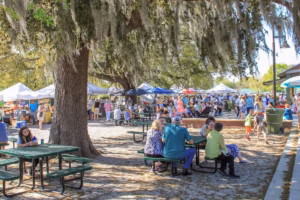 Outdoor farmers market with vendor tents and families sitting at green picnic tables beneath large trees.