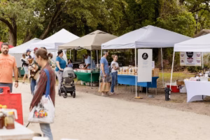 Outdoor farmers market stalls with white canopy tents along a sandy path in a park, with shoppers and vendors browsing and a dog being carried past the booths.