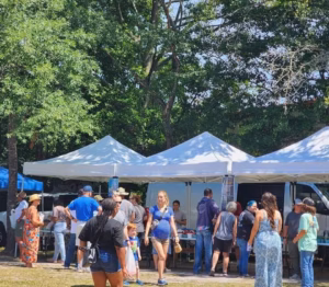 People browse under white canopy tents at the Ridgeville Farmers Market in a sunny outdoor setting with tall green trees.