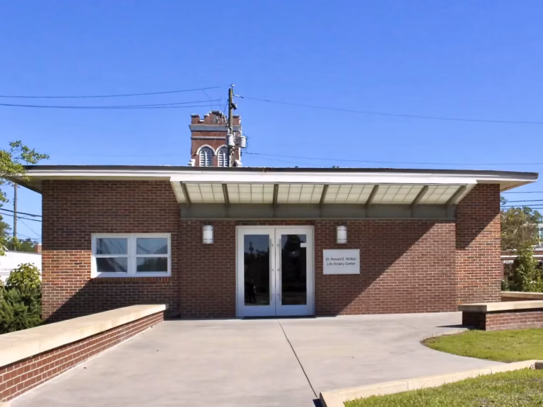 Front entrance of the Dr. Ronald E. McNair Life History Center with brick facade, arched brick details, and a concrete walkway under a clear blue sky.