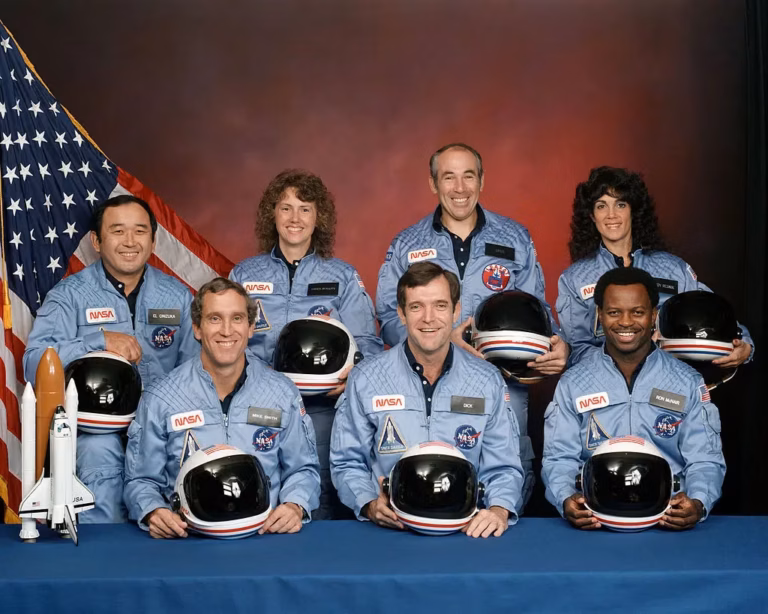NASA shuttle crew portrait with seven astronauts in light-blue flight suits holding black helmets and posing beside a small rocket model in front of a U.S. flag.