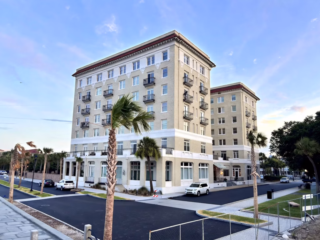 Exterior view of a multi-story building with light stone walls, white railings, small black balcony railings, and red trim, with green fronds of Palmetto trees and a few cars parked along the blacktop street.