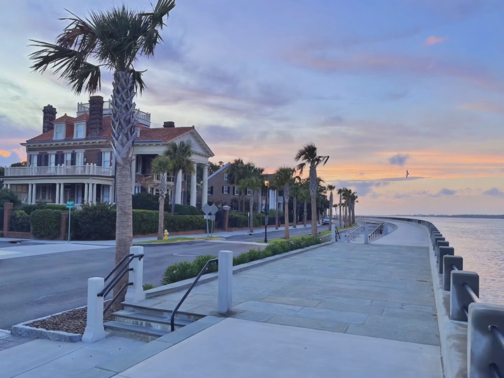 Sunrise at Murray Boulevard along a waterfront walkway, with palm trees and white railings leading to the sea beside a two-story brick home with columns.